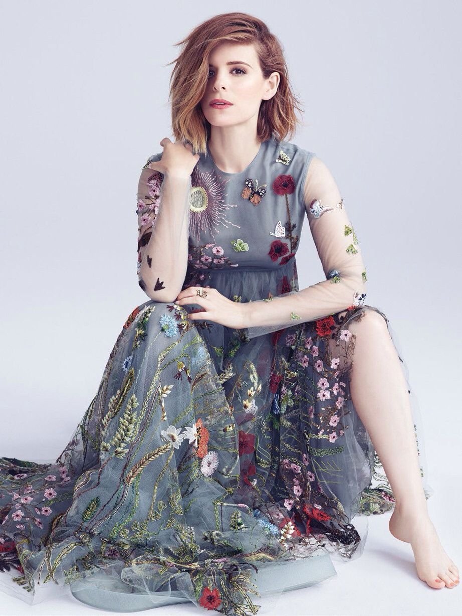 Adult female with light brown hair sits in a sheer, long-sleeved, floral-embroidered gown, gazing forward in a brightly lit indoor photo studio setting.