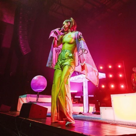 A female singer stands on a stage with colorful lights, wearing green pants and singing into a microphone during a musical performance.