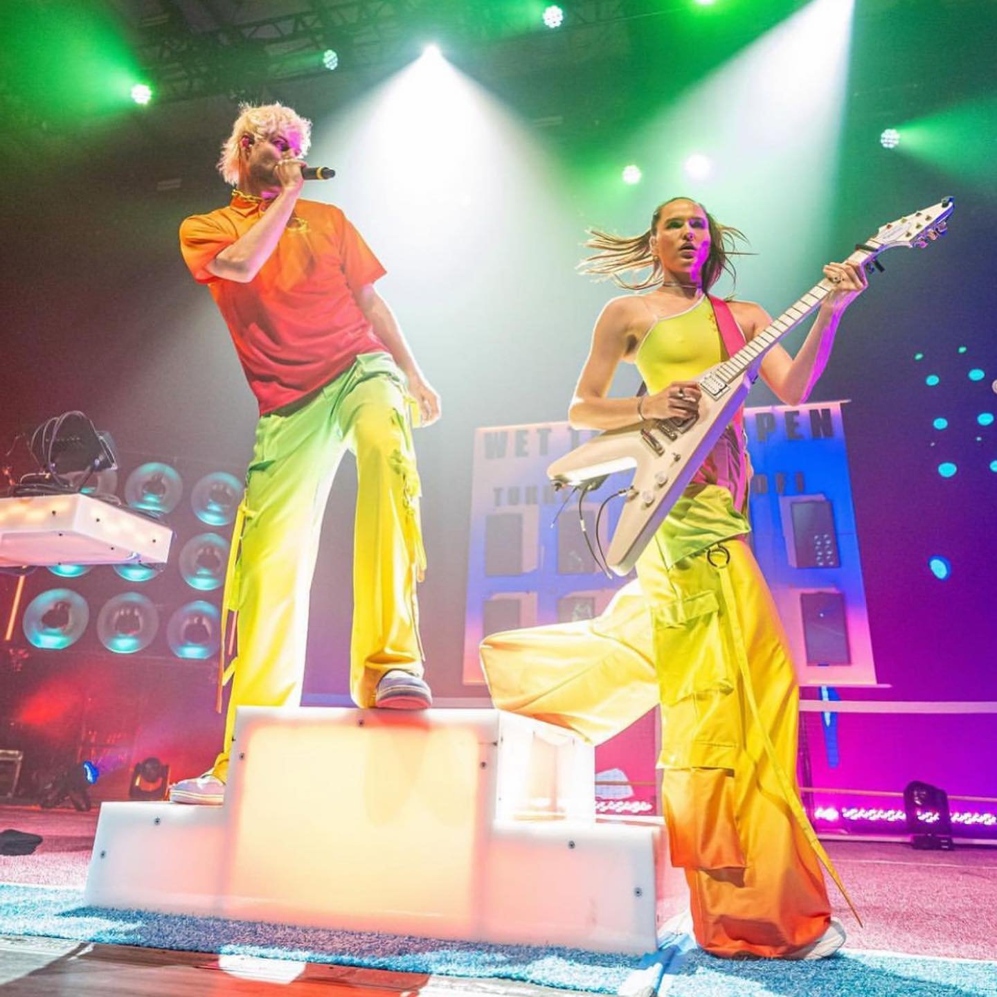 Two musicians perform on a brightly lit stage, one singing into a microphone and the other playing an electric guitar, creating a vibrant scene.