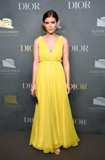 Woman in a yellow gown attends the Guggenheim International Gala with a gold clutch, captured in a medium shot on a gray branded backdrop.