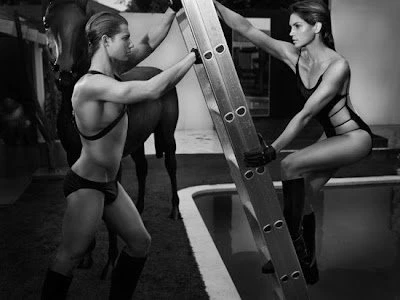 Two adult figures in swimwear and boots pose with a ladder and horse in a studio setting, creating a high-contrast black and white image.