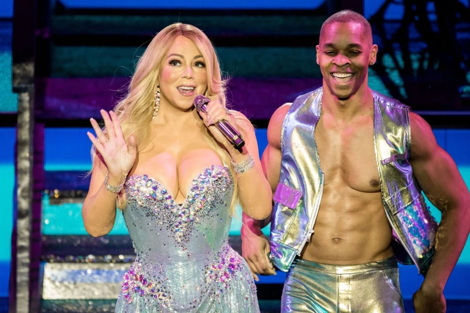 Mariah Carey performs on stage with a male dancer, both in sparkling costumes, under bright stage lighting and a blue background.