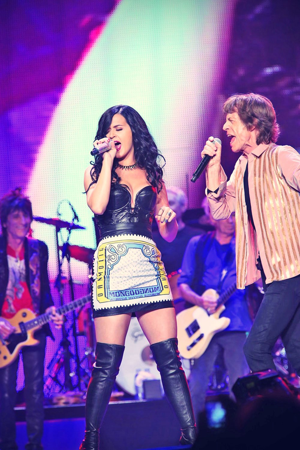 Katy Perry and Mick Jagger perform on stage at a concert with other musicians, bright stage lights, and a crowd in the background.