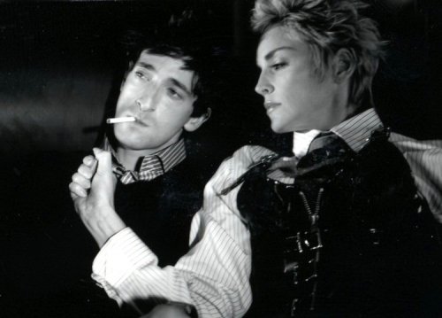 Monochrome image of a man smoking and a woman in a black top with short blonde hair, posing in front of a dark background.