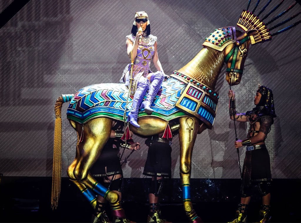 Singer Katy Perry performs on a gold-colored horse statue, wearing a purple outfit with an Egyptian-inspired theme during a concert performance.