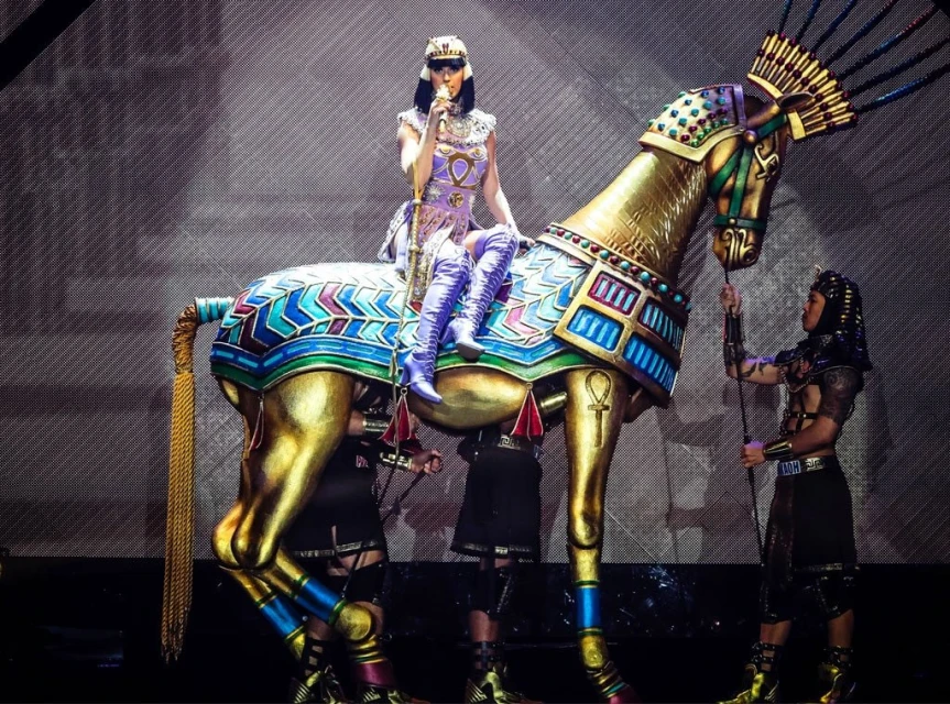 Singer Katy Perry performs on a gold-colored horse statue, wearing a purple outfit with an Egyptian-inspired theme during a concert performance.