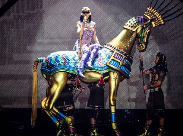 Singer Katy Perry performs on a gold-colored horse statue, wearing a purple outfit with an Egyptian-inspired theme during a concert performance.