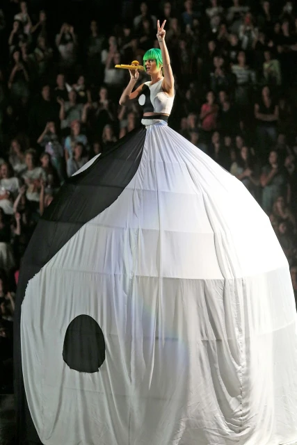 Singer performs on stage wearing a large yin and yang costume, holding a microphone, with crowd in the background.