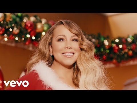 Close-up shot of Mariah Carey wearing a red and white Santa-themed outfit in front of a decorated Christmas tree.