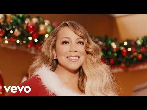 Close-up shot of Mariah Carey wearing a red and white Santa-themed outfit in front of a decorated Christmas tree.