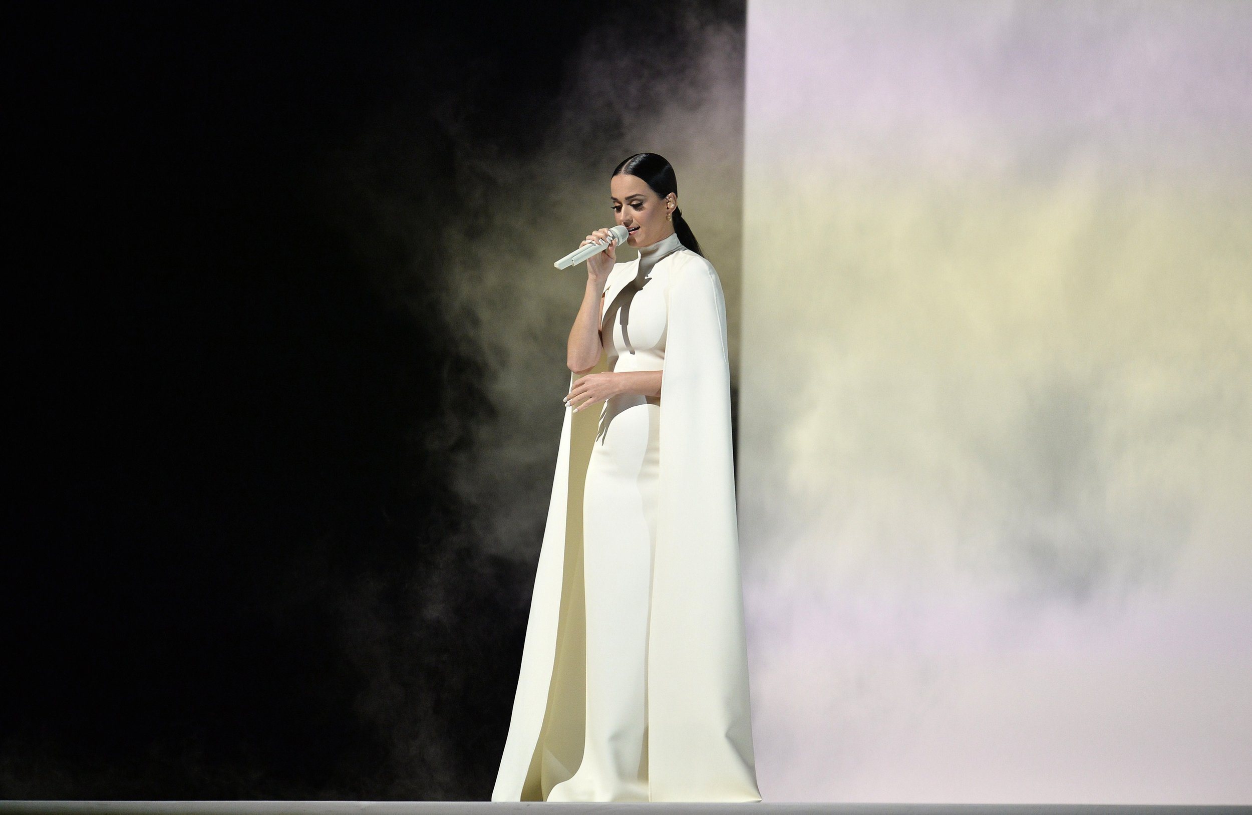Woman in white outfit sings with microphone against a backdrop of black and white smoke, captured in a medium shot.