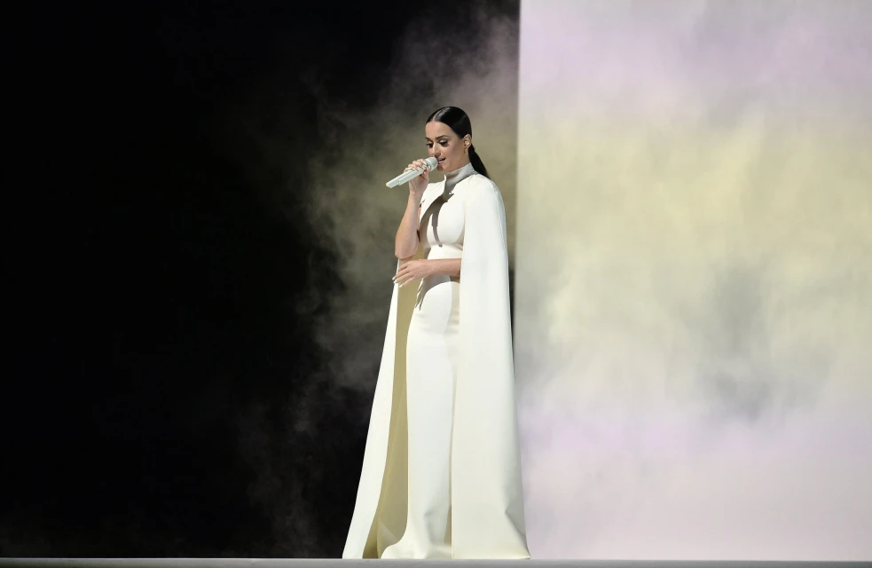 Woman in white outfit sings with microphone against a backdrop of black and white smoke, captured in a medium shot.