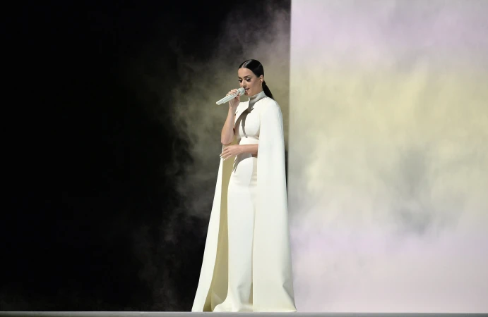 Woman in white outfit sings with microphone against a backdrop of black and white smoke, captured in a medium shot.