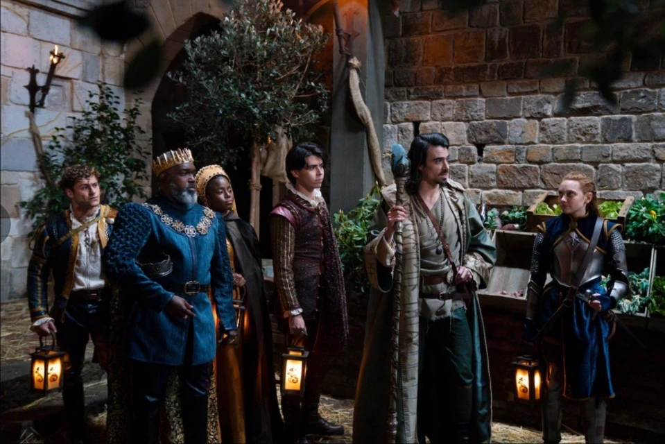 Six people in medieval-style costumes stand outdoors by a stone wall, with lanterns and props, illuminated by ambient and lantern light.