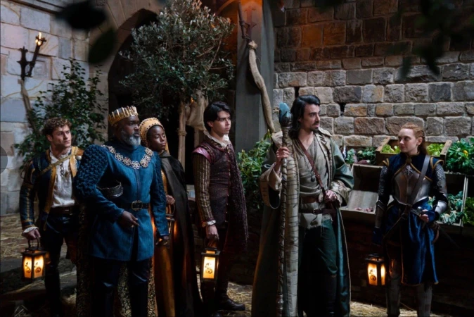 Group of six people in period costumes standing in front of a stone wall. One man wears a crown; the others carry props and lanterns.