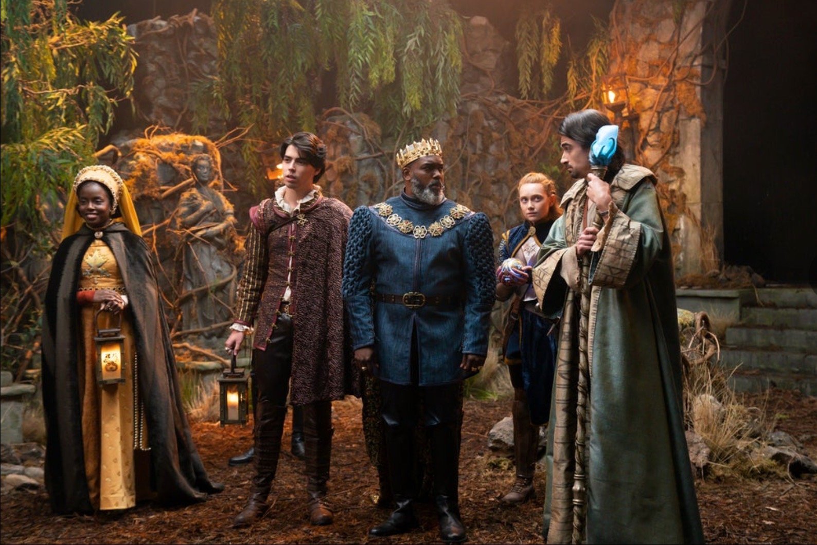 Group of five people in fantasy attire standing outdoors amidst trees and foliage, featuring regal robes, crowns, and medieval elements.