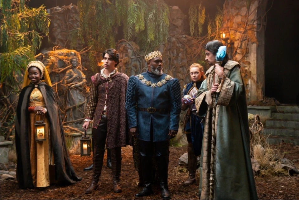 Group of five people in fantasy attire standing outdoors amidst trees and foliage, featuring regal robes, crowns, and medieval elements.