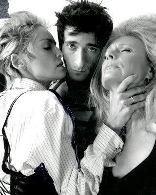 Black and white studio shot of actor Adrien Brody between two light-haired women in dress shirts, one with hand at his neck.