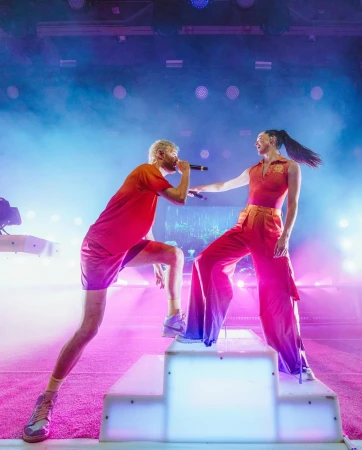 Two musicians, a man and a woman, perform on stage in red outfits under bright lights, singing into microphones on a tiered platform.