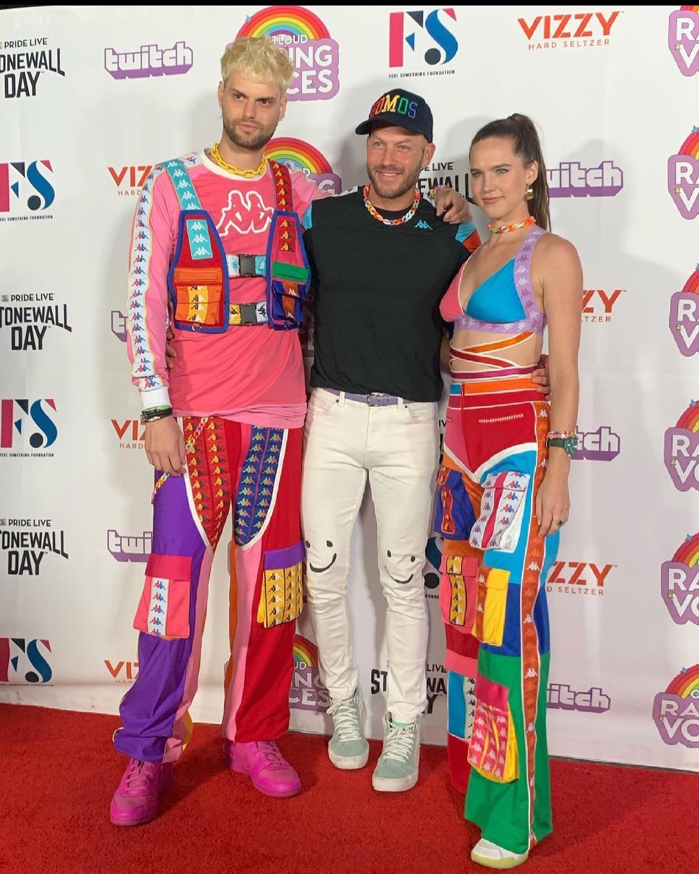 Three adults pose on a red carpet at a Pride event, showcasing bright, patchwork-style clothing and a colorful, celebratory atmosphere.