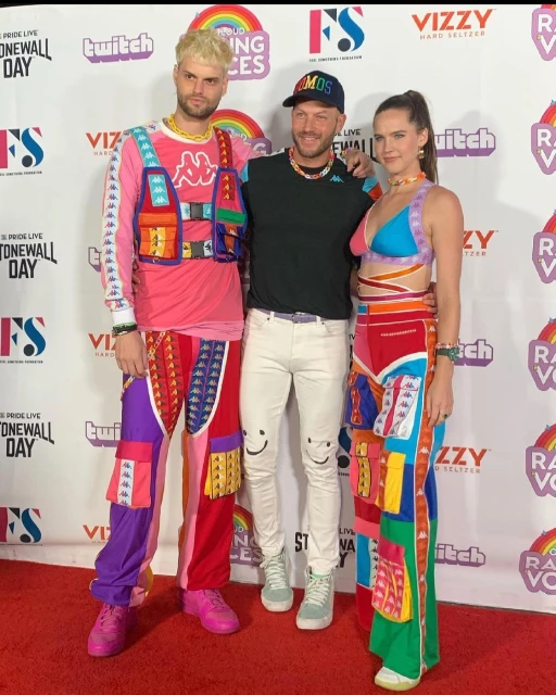 Three adults pose on a red carpet at a Pride event, showcasing bright, patchwork-style clothing and a colorful, celebratory atmosphere.