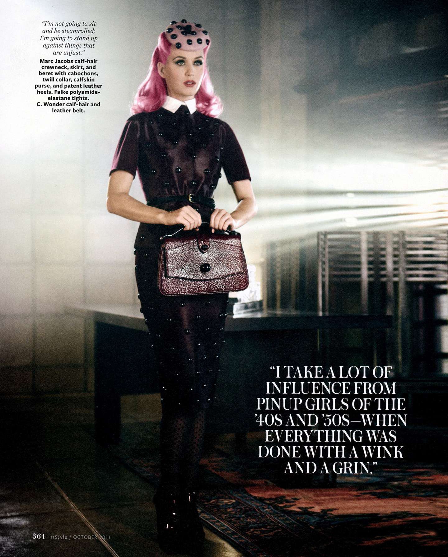 Full-length portrait of a woman with pink hair in a dark studded dress and hat holding a purse in a studio environment.