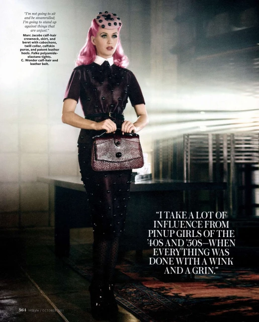 Full-length portrait of a woman with pink hair in a dark studded dress and hat holding a purse in a studio environment.