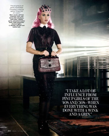Full-length portrait of a woman with pink hair in a dark studded dress and hat holding a purse in a studio environment.