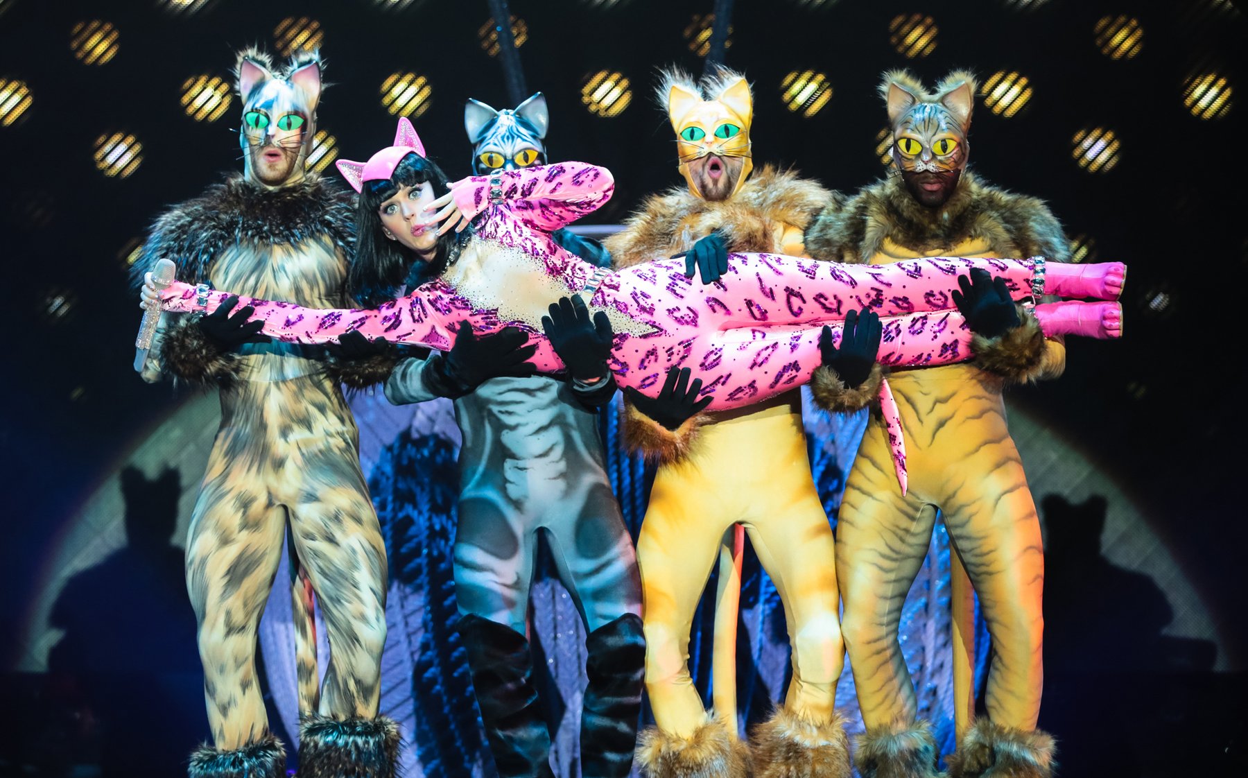 Five performers in cat costumes on stage, woman carried by four men, illuminated by stage lights, with a patterned background.