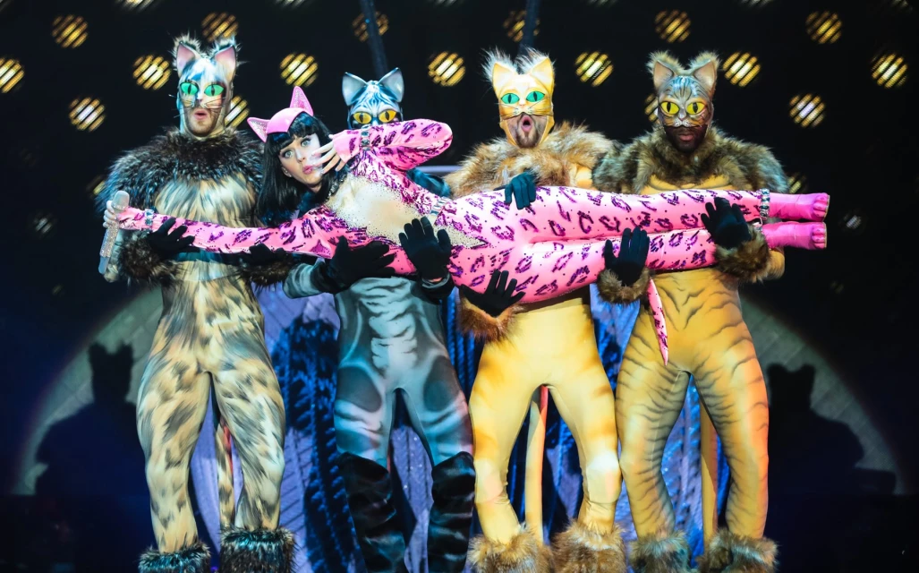 Five performers in cat costumes on stage, woman carried by four men, illuminated by stage lights, with a patterned background.