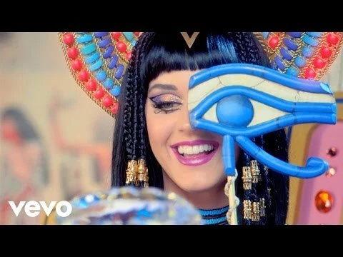 Close-up of a smiling woman with Egyptian-style makeup and braids, wearing a colorful beaded headdress and holding an ankh.