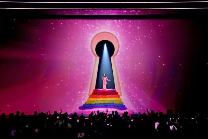 Female singer in pink dress on rainbow platform in front of keyhole-shaped screen and crowd on stage at a music concert.