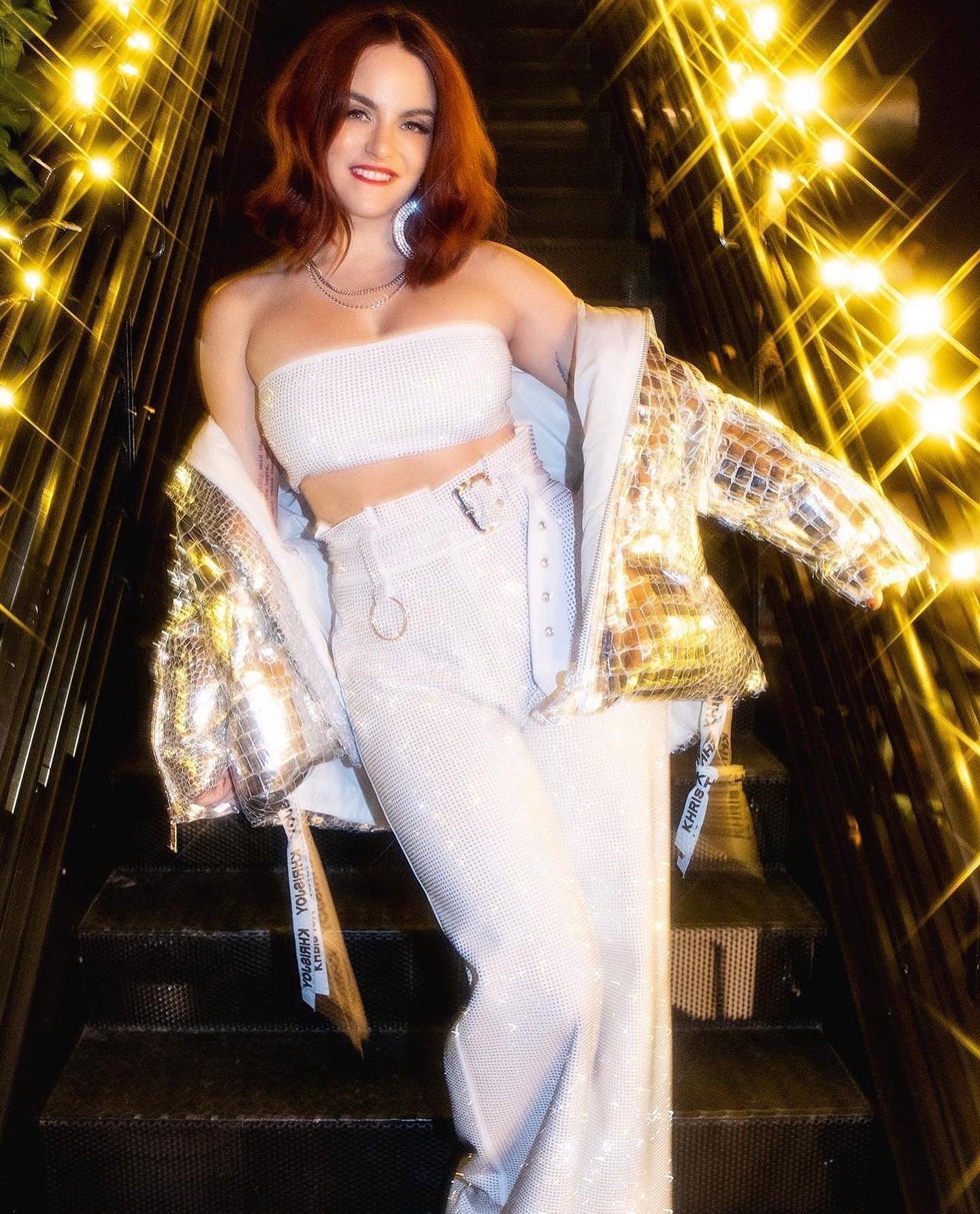 A smiling woman with auburn hair poses on stairs in a matching white tube top and pants, adorned with a gold mirror jacket and gold lights.