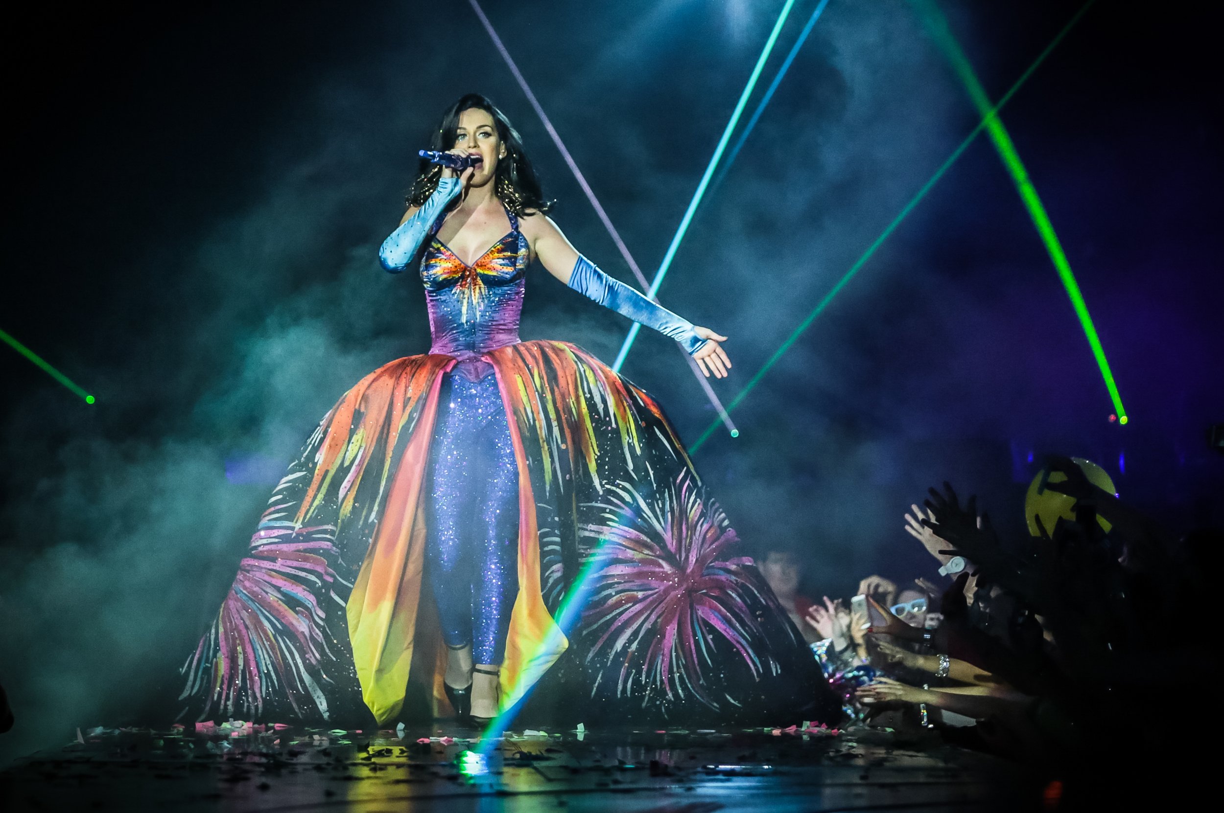 A singer performs on stage in a colorful gown, singing into a microphone, with stage lights and a crowd visible in the background.