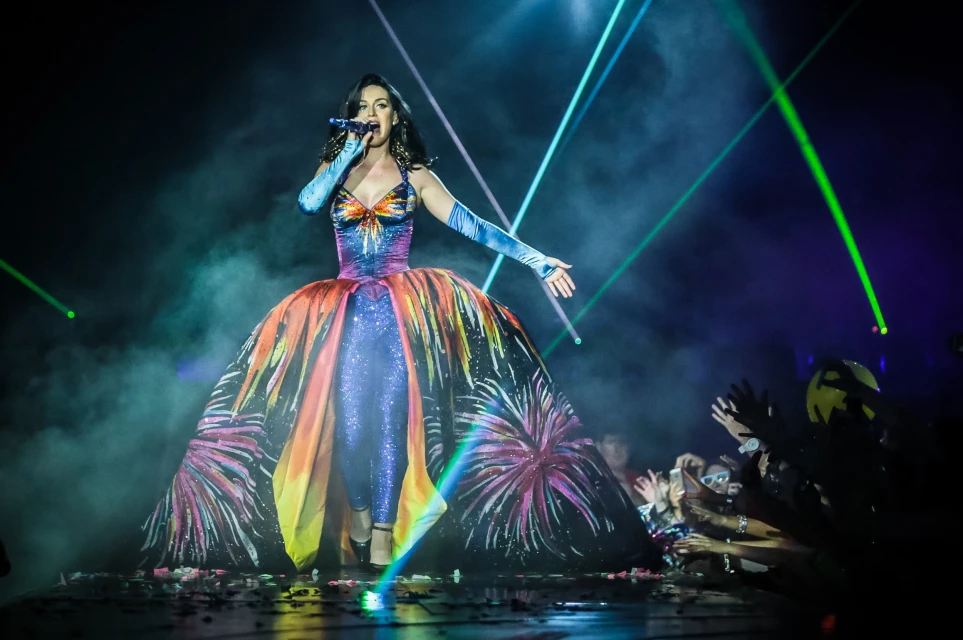 A singer performs on stage in a colorful gown, singing into a microphone, with stage lights and a crowd visible in the background.