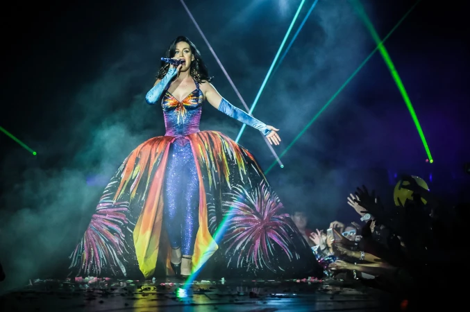 A singer performs on stage in a colorful gown, singing into a microphone, with stage lights and a crowd visible in the background.