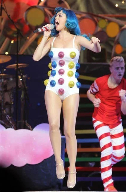 Katy Perry performs live on stage, wearing a white bodysuit with colorful polka dots and blue hair, singing into a microphone.