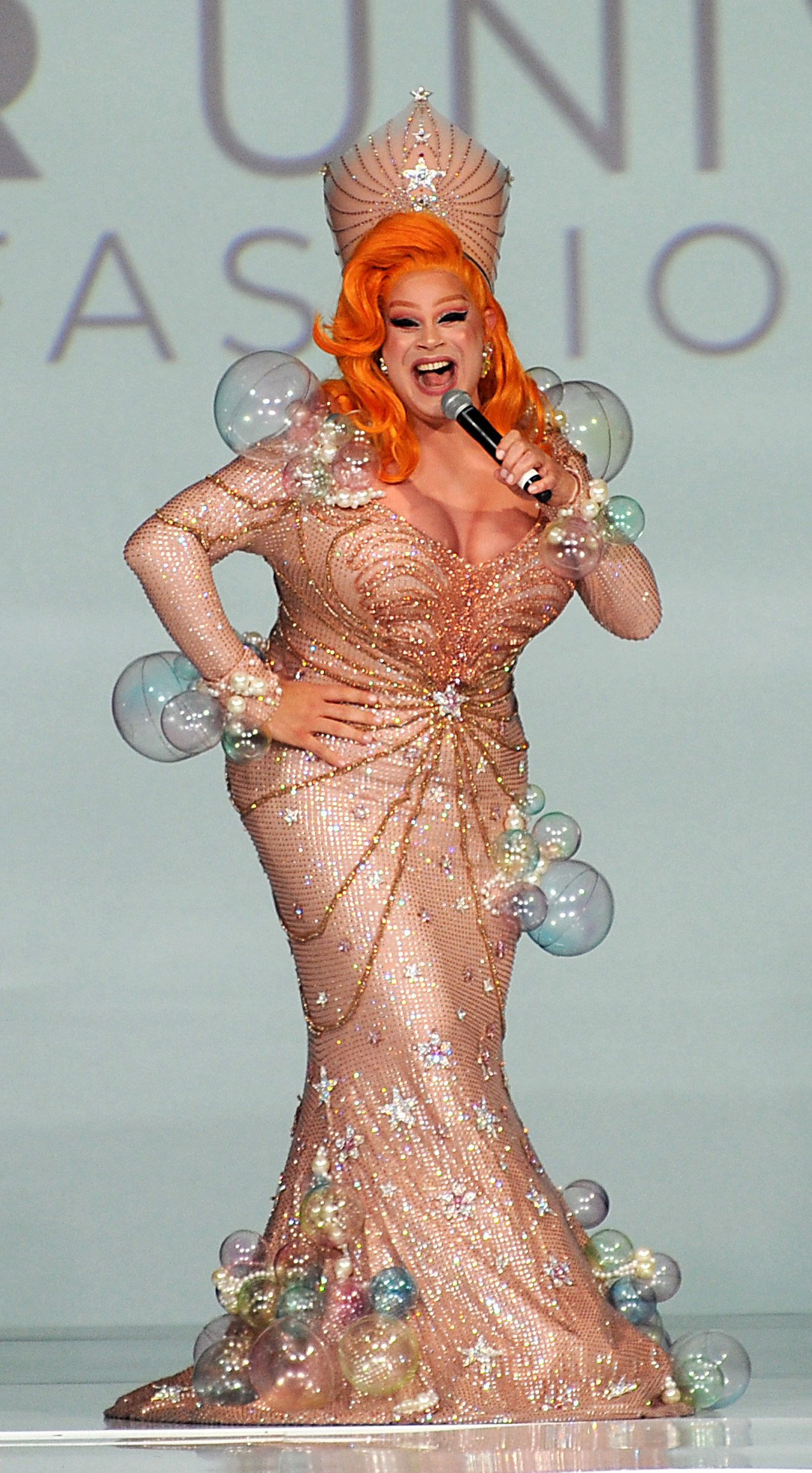 Female performer in sparkly gown on runway, holding a microphone with orange hair and wearing a crown on stage during a fashion show.
