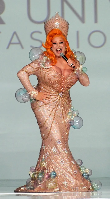 Female performer in sparkly gown on runway, holding a microphone with orange hair and wearing a crown on stage during a fashion show.