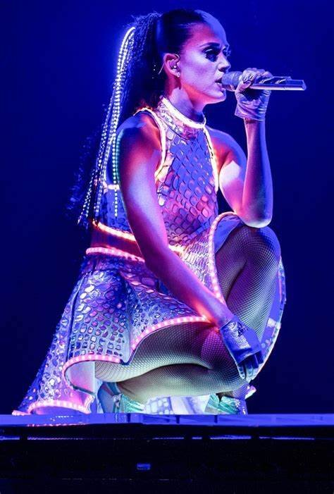 Singer Katy Perry performs in a futuristic outfit with light-up details while crouching on stage and holding a microphone in a blue-lit concert.