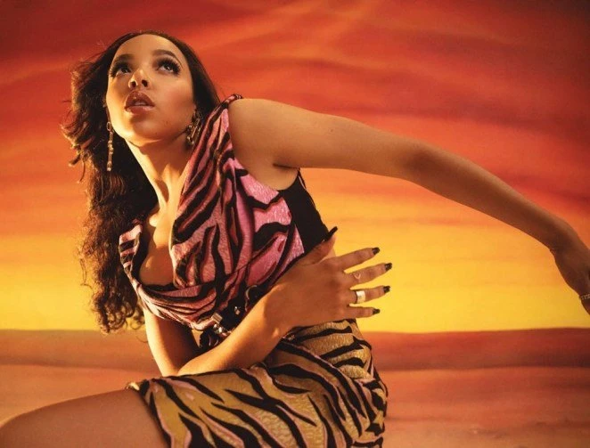 A woman in a pink and tan animal print top and skirt poses against an orange and yellow background in a fashionable pose.