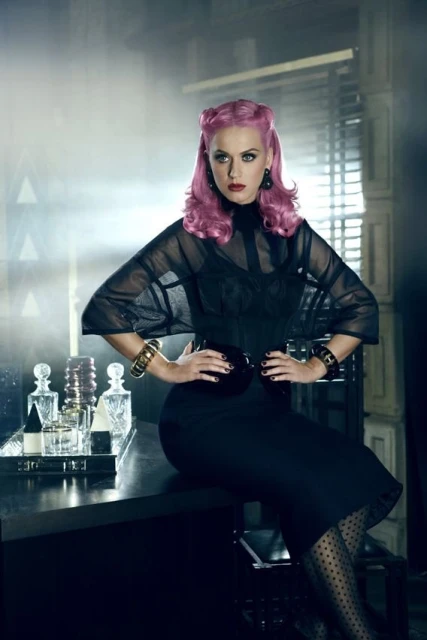 Glamorous woman with pink hair poses at a bar with her hands on her hips. She wears a sheer black top, a black pencil skirt, and gold bracelets.