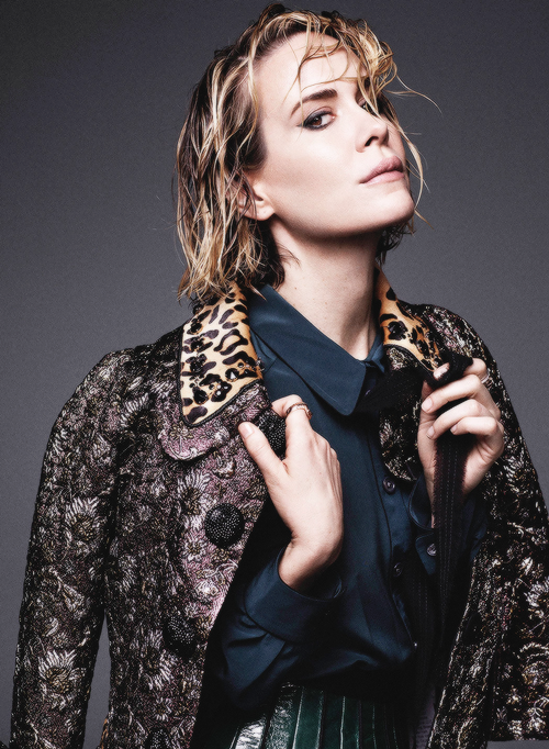Stylish portrait of a blonde woman in a patterned coat and teal shirt, shot in a photo studio against a gray backdrop.