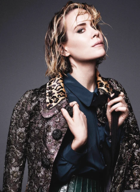 Stylish portrait of a blonde woman in a patterned coat and teal shirt, shot in a photo studio against a gray backdrop.