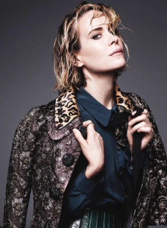 Stylish portrait of a blonde woman in a patterned coat and teal shirt, shot in a photo studio against a gray backdrop.