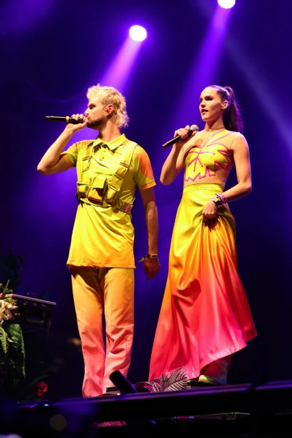 Two singers perform on stage with microphones, wearing colorful yellow and orange outfits, illuminated by bright stage lighting.