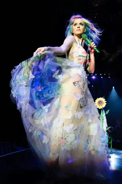 Female singer on stage in butterfly gown with multicolor hair, holding microphone during music performance in dark, dynamic lighting.