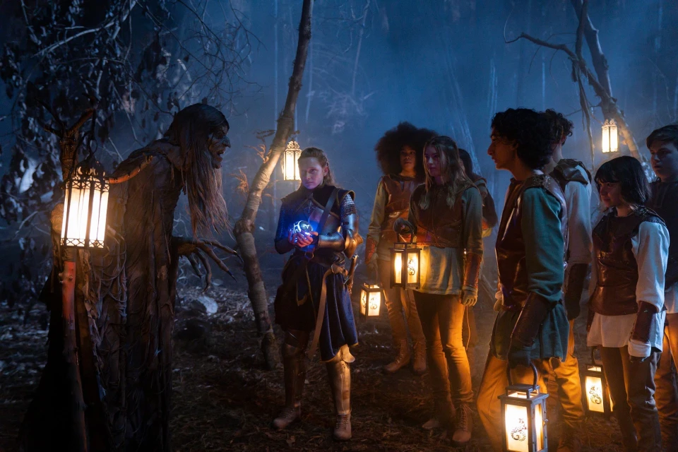 A group of young people, some holding lanterns, stand in a misty forest with a tree-like creature in a dimly lit scene.