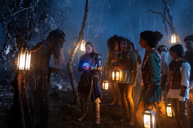 A group of young people, some holding lanterns, stand in a misty forest with a tree-like creature in a dimly lit scene.