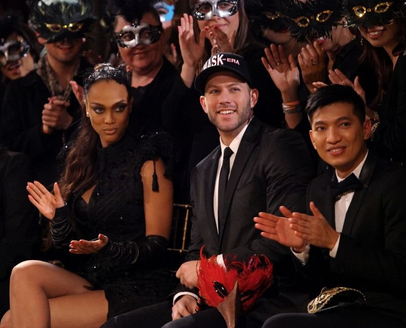 Tyra Banks and two other seated guests wearing formal wear are clapping at a fashion show with an audience in the background.